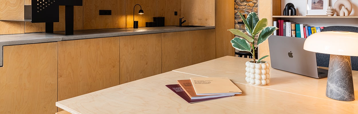 Un espace de travail moderne avec une table en bois, un MacBook, des livres et une plante dans un vase texturé. En arrière-plan, on trouve une cuisine minimaliste avec des meubles en bois, un robinet noir et une petite lampe de table offrant un éclairage d'ambiance.