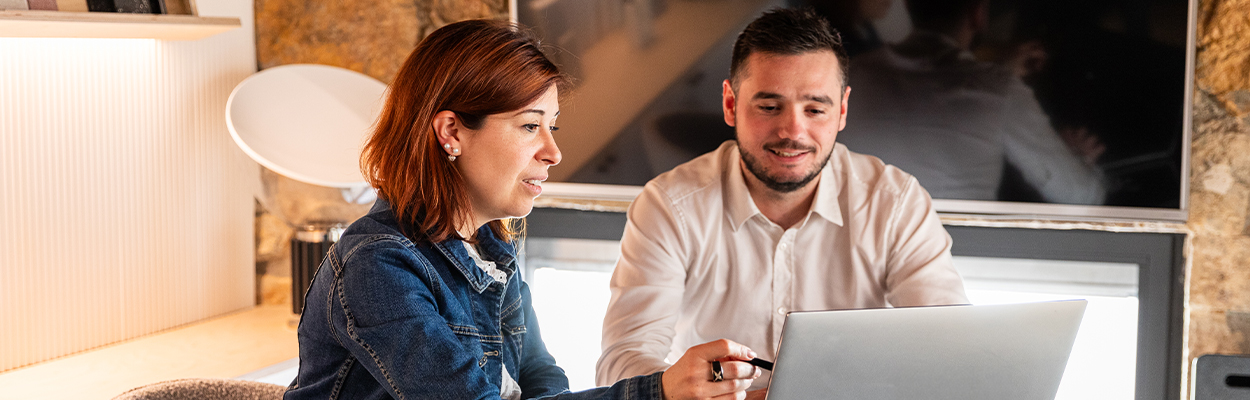 Deux personnes, l'une vêtue d'une veste en jean et l'autre d'une chemise blanche, sont assises à un bureau et travaillent sur un ordinateur portable. Elles semblent concentrées et engagées dans une discussion. L'arrière-plan comprend un intérieur moderne et confortable avec un écran de télévision et des éléments muraux en pierre.