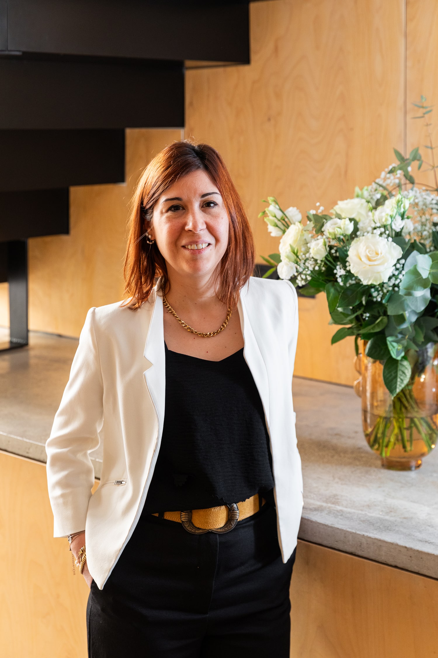 Une personne aux cheveux mi-longs sourit tout en portant un blazer blanc sur un haut et un pantalon noirs. Elle se tient debout à côté d'un comptoir en béton avec un bouquet décoratif de fleurs blanches dans un vase en verre. L'arrière-plan est constitué de panneaux de bois et d'un escalier noir.