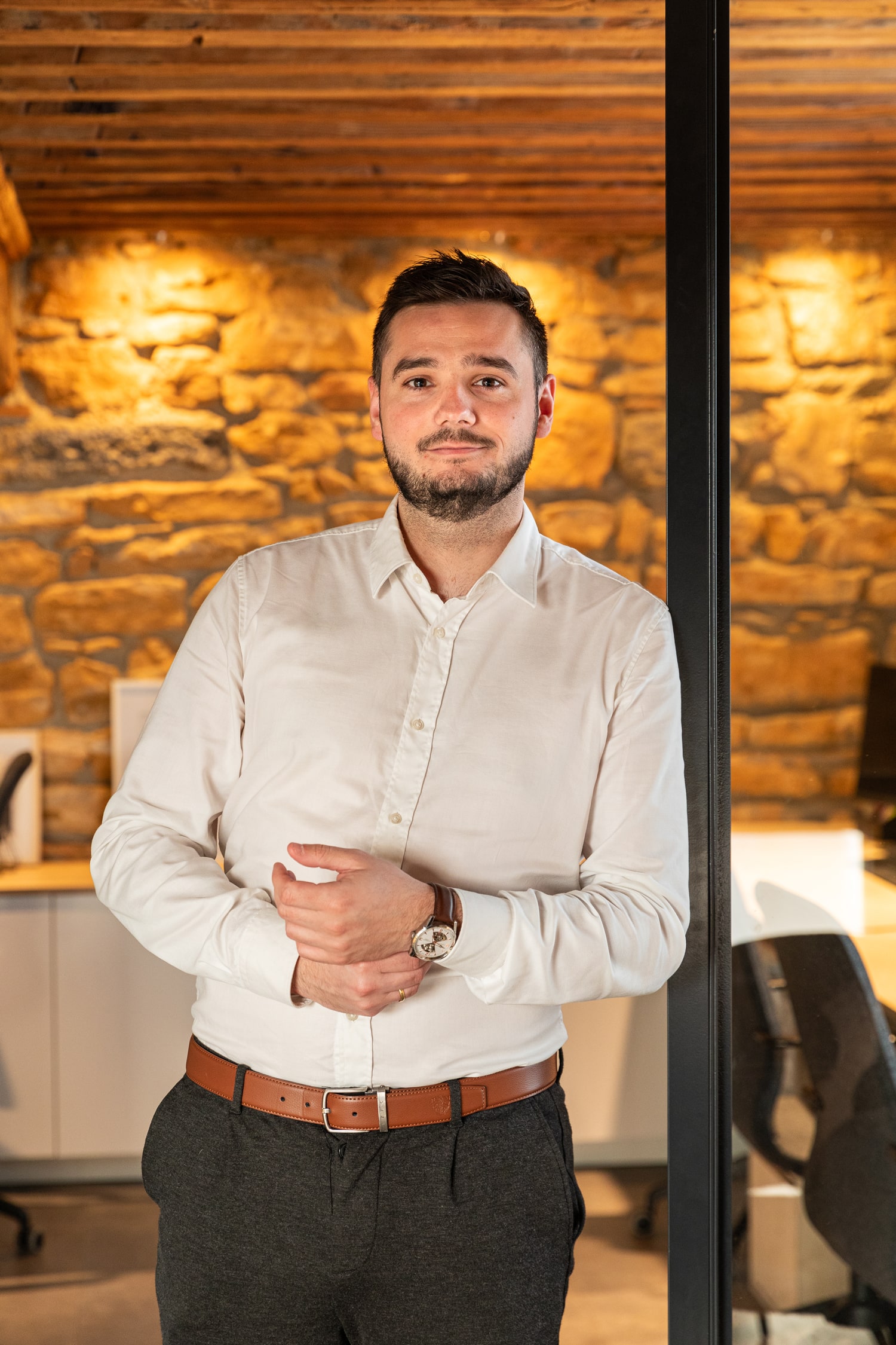 Un homme aux cheveux noirs courts et à la barbe, vêtu d'une chemise blanche boutonnée et d'un pantalon foncé, se tient debout, les mains jointes devant lui. Il se trouve dans un espace de bureau moderne avec un éclairage chaleureux et un mur de pierre en arrière-plan.