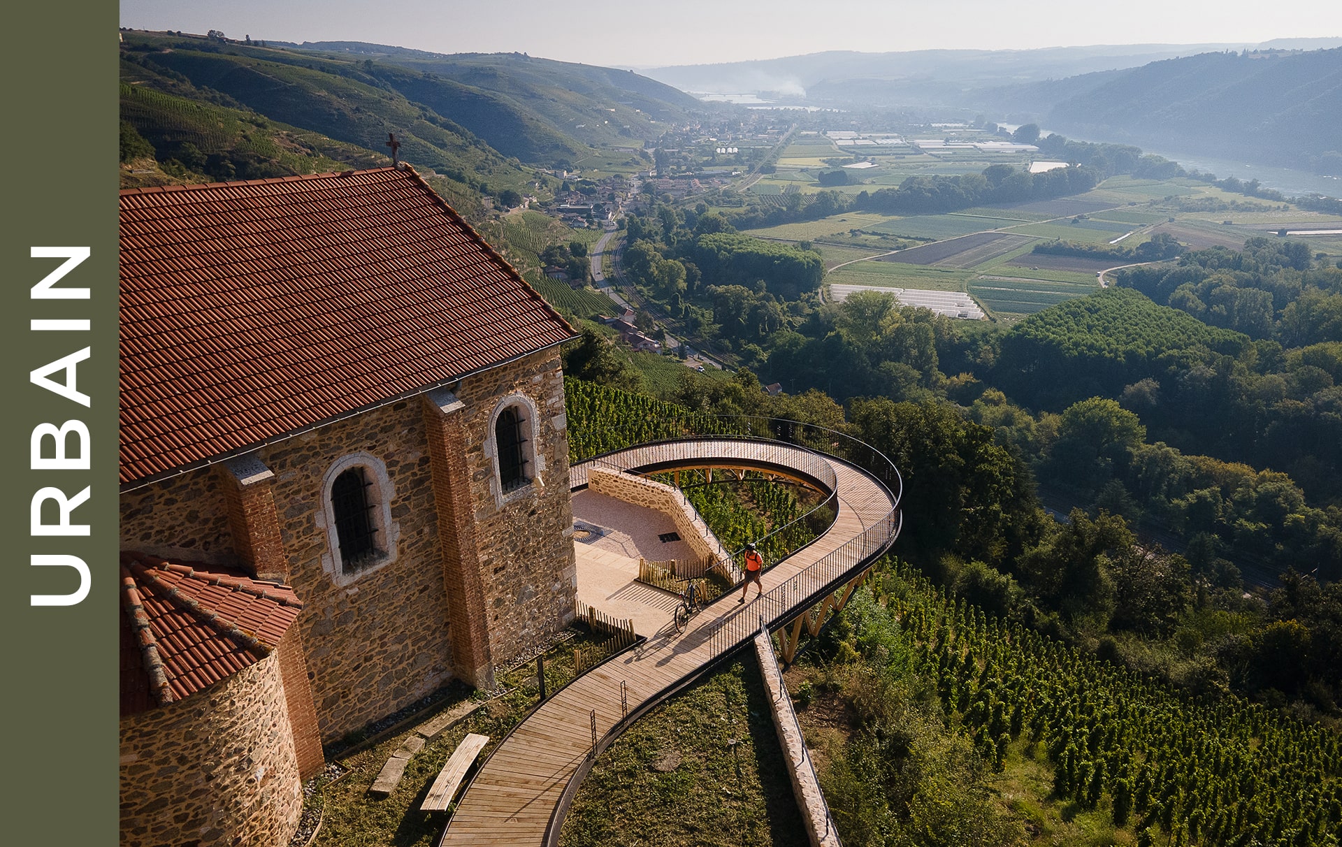 Vue panoramique d'un bâtiment rustique en pierre avec un toit en terre cuite dans un paysage de vignobles vallonnés. Une passerelle en bois sinueuse mène à la structure, entourée d'une végétation luxuriante et surplombant une vallée avec de petites villes et des montagnes lointaines.