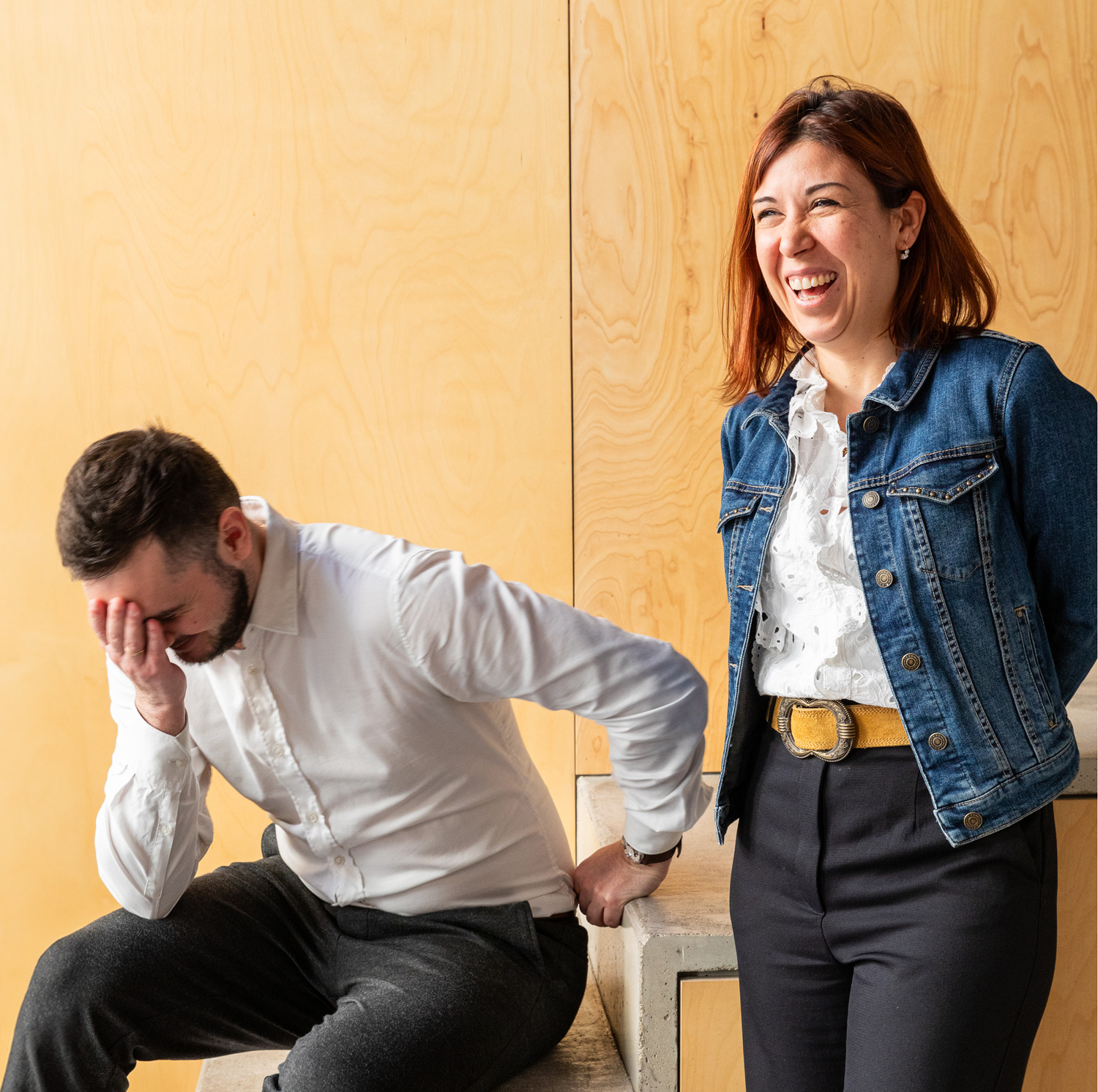 Un homme et une femme partagent un moment de détente. L'homme, vêtu d'une chemise blanche et d'un pantalon sombre, se couvre le visage d'une main alors qu'il est assis sur des marches en béton. La femme se tient à côté de lui, vêtue d'une veste en jean et rigole, avec des panneaux en bois en arrière-plan.